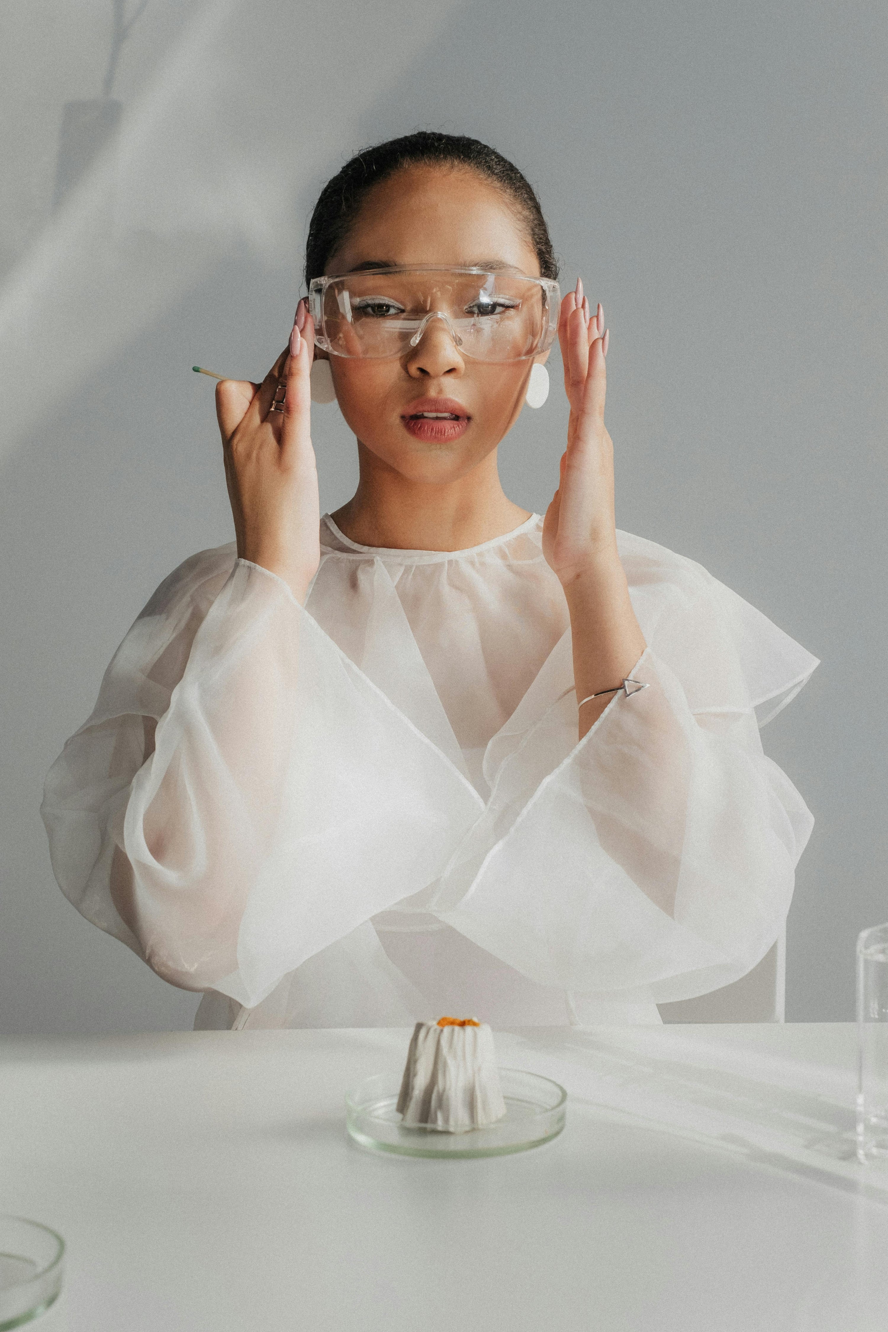 Woman in a sheer white blouse and safety goggles seated at a white table with a small sample in a petri dish, evoking a clean, scientific beauty lab aesthetic.