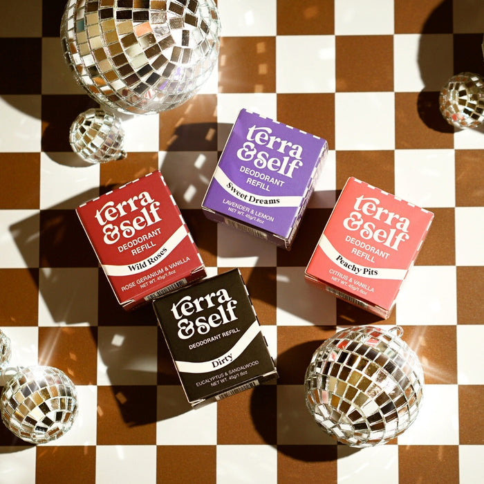 Top-down view of all four Terra & Self plastic-free deodorant refill boxes — Wild Roses (red), Sweet Dreams (purple), Peachy Pits (orange), and Dirty (black) — arranged on a brown and white checkered surface surrounded by mini disco balls casting light reflections.