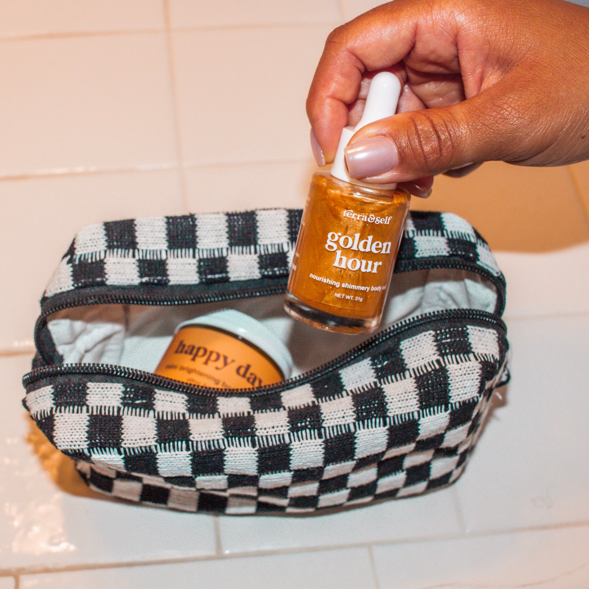 Golden Mini Body Oil Bottle being pulled out of checkered makeup bag on counter